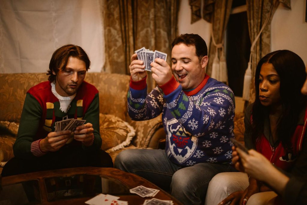 Group of friends in cozy sweaters laughing and playing cards indoors during a Christmas party.