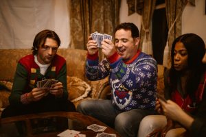Group of friends in cozy sweaters laughing and playing cards indoors during a Christmas party.